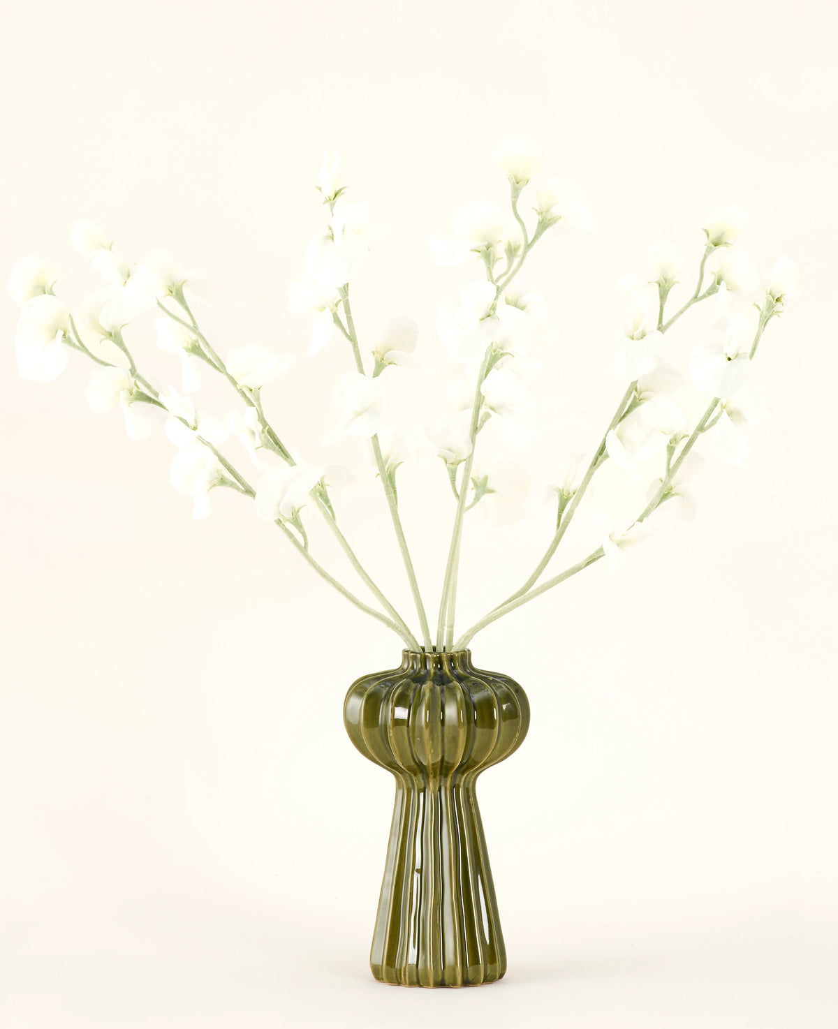 Un vase en verre nervuré vert contient le bouquet de fleurs artificielles en soie Crème de pois avec de délicates fleurs en soie blanche et des tiges minces, sur un fond uni blanc cassé.