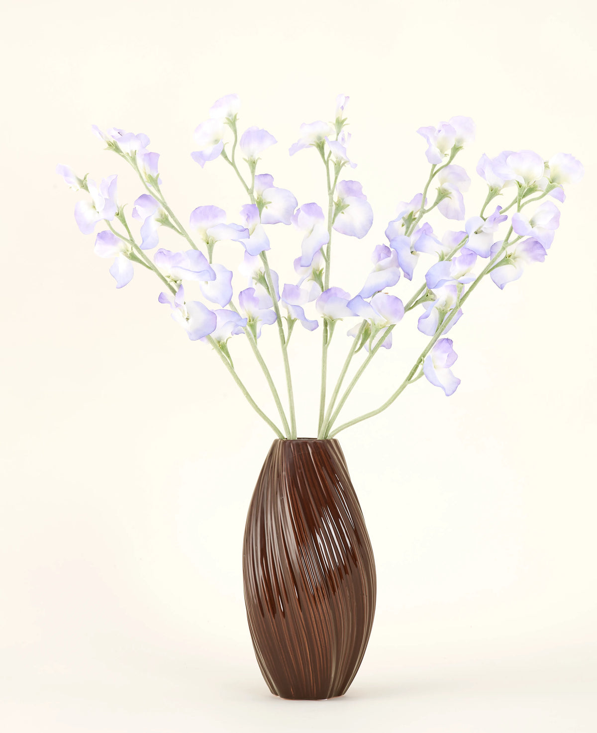 Un vase marron nervuré contient le bouquet de fleurs artificielles en soie, pois lavande, avec des pétales délicats et des tiges vertes sur un fond blanc cassé uni pour une atmosphère subtile de cottage.