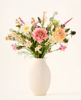 Dans un vase en céramique blanche se trouve le bouquet de fleurs artificielles en soie « Backyard Blossom », composé d'un bel arrangement de roses roses, de marguerites blanches et de petites fleurs jaunes, faites de fleurs en soie, complétées par des feuilles vertes luxuriantes sur un fond clair uni.