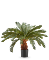 La Plante Artificielle Cycas UV 90 cm possède de longues feuilles vertes courbées et une tige brune épaisse dans un pot noir, sur un fond blanc uni.
