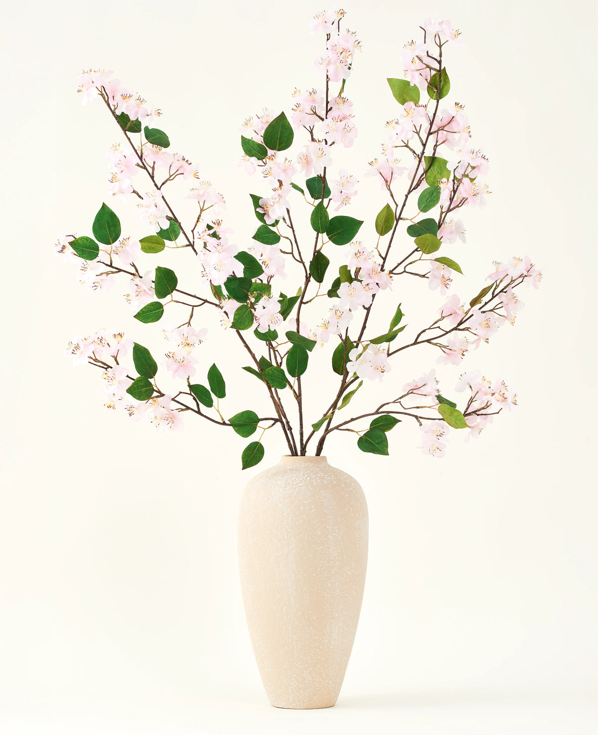 Un vase en céramique beige contient plusieurs longues branches avec le bouquet de fleurs artificielles en soie de feuilles roses et vertes de fleurs de pêcher, sur un fond blanc uni - parfait pour un intérieur floral artificiel.