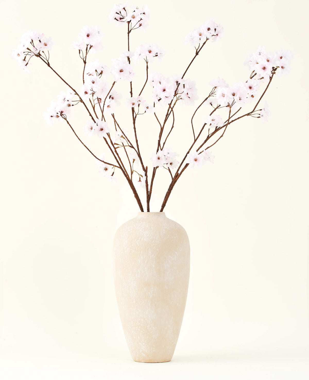 Le bouquet de fleurs artificielles en soie Fleur de cerisier rose clair 114 cm se dresse dans un grand vase en céramique beige avec de longues et fines branches et de délicates fleurs rose clair, créant un accent floral artificiel serein sur un fond uni.