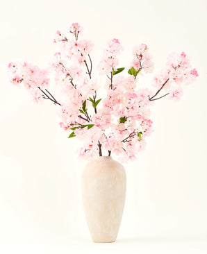 Le bouquet de fleurs artificielles en soie Fleur de cerisier rose 119 cm, disposé dans un vase en céramique beige aux délicates feuilles vertes, crée un accent intérieur serein sur un fond clair simple.