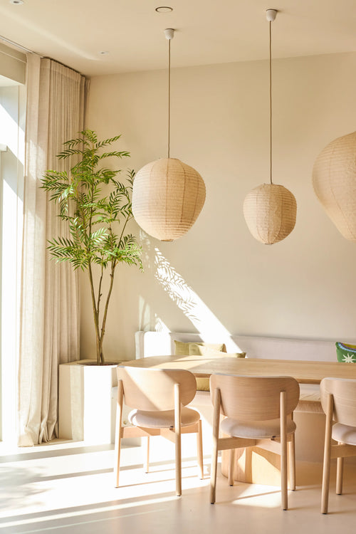 Une salle à manger lumineuse et minimaliste est meublée de chaises en bois clair, d'une table, de trois lampes suspendues en papier, de la plante artificielle Mahonia de 210 cm en pot et de rideaux allant du sol au plafond près d'une grande fenêtre laissant entrer la lumière du soleil.