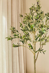 Un olivier artificiel de 150 cm, décoré d'olives foncées, est attrayant à l'intérieur à côté de rideaux beiges et d'un mur marron clair, et donne un aspect calme et naturel - idéal pour tout intérieur avec un olivier.