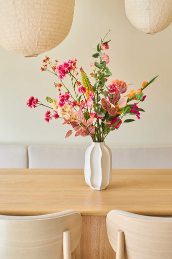 Sur une table en bois clair se trouve un vase blanc avec un bouquet de "Fleurs artificielles en soie Pêche Sauvage", composé de fleurs d'été. Deux chaises en bois clair et de grandes lampes suspendues en papier complètent l'intérieur minimaliste et cosy.
