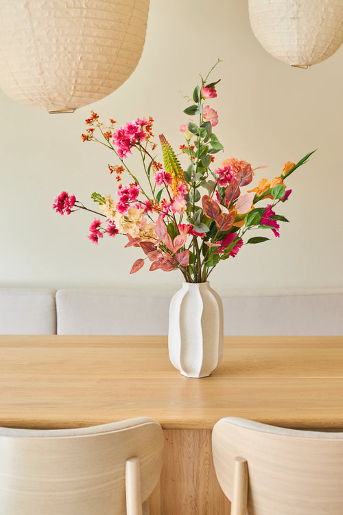 Sur une table en bois clair se trouve un vase blanc avec un bouquet de "Fleurs artificielles en soie Pêche Sauvage", composé de fleurs d'été. Deux chaises en bois clair et de grandes lampes suspendues en papier complètent l'intérieur minimaliste et cosy.