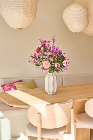 Sur une table à manger en bois clair se trouve un vase blanc avec le bouquet de fleurs artificielles en soie Purple Dawn, débordant de teintes roses et violettes vibrantes. Derrière lui, un banc rembourré avec des coussins et deux lanternes en papier suspendues complètent la scène, imprégnant l'espace de l'atmosphère tranquille inspirée de Purple Dawn.