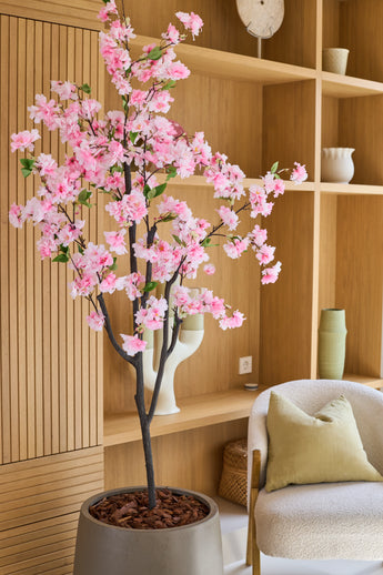 La Fleur de Cerisier Artificielle 150 cm rose, avec ses délicates fleurs en soie rose, sera élégante dans une pièce moderne avec des étagères en bois, des objets décoratifs, une chaise beige et un coussin vert – ajoutant le charme des fleurs en soie à votre intérieur.