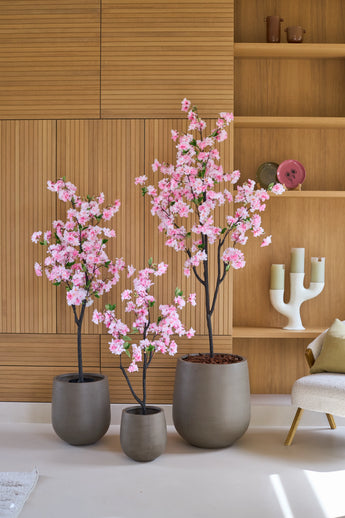 Trois cerisiers artificiels de 150 cm aux fleurs roses se dressent devant un lambris en bois, à côté d'étagères décorées de décorations et d'un bougeoir blanc moderne.