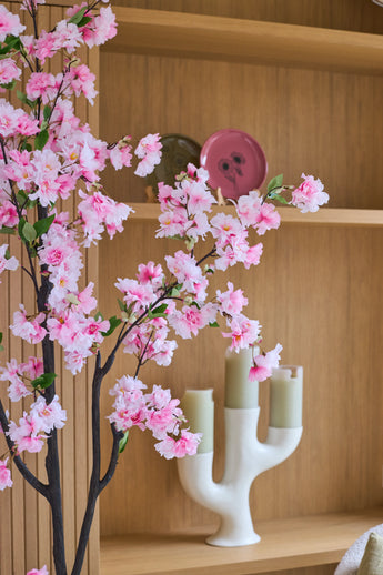 La « Fleur de cerisier artificielle » (150 cm) aux branches roses est placée devant des étagères en bois, sur lesquelles sont disposées des bougies vertes dans un chandelier blanc et des assiettes décoratives.
