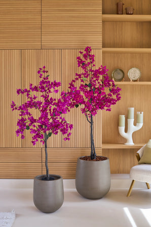 Deux plantes en pot de type bougainvillier artificiel de 180 cm, de couleur violet, avec des fleurs violettes vibrantes se dressent devant un mur en bois clair et des étagères avec des objets décoratifs tels que des bougies et des assiettes. À droite, une chaise beige, qui ajoute une ambiance cosy à ce salon végétal artificiel animé.