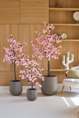 Trois bougainvilliers artificiels de 180 cm, de couleur rose et avec des fleurs roses, se dressent devant des étagères murales en bois, à côté d'une chaise blanche moderne avec un coussin.