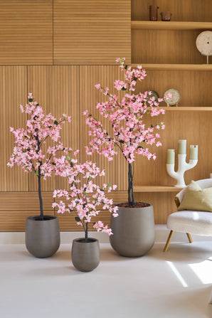 Trois plantes en pot de type Plante Artificielle Bougainvillier de 120 cm, de couleur rose et à fleurs roses, se trouvent sur un sol blanc dans une pièce moderne avec des boiseries, des étagères et une chaise blanche.