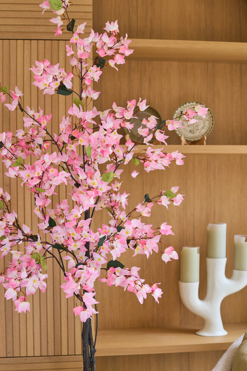Un bougainvillier artificiel de 150 cm, de couleur rose et avec des fleurs roses, se dresse devant des étagères en bois sur lesquelles sont exposés une assiette décorative et un bougeoir blanc moderne avec trois bougies vertes.