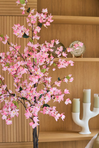 Devant les étagères en bois, sur lesquelles sont disposés une assiette et un chandelier blanc avec trois bougies vertes, se trouve une plante artificielle de Bougainvillier rose de 120 cm.