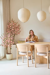Une femme aux longs cheveux blonds est assise à une table en bois clair, lisant un livre et souriant. À côté d'elle se trouve la plante de cerisier artificielle de 150 cm rose, dont les fleurs roses apportent un accent vivant. Les lampes suspendues et la décoration minimaliste créent une atmosphère légère et cosy.