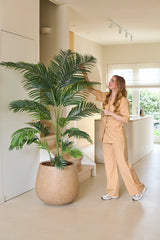 Une femme en costume beige se tient à l'intérieur, souriante, tout en touchant les feuilles de la plante artificielle Phoenix 200 cm, près d'un escalier dans une pièce lumineuse et moderne.
