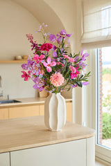 Sur un îlot de cuisine en bois clair se trouve un vase blanc avec le bouquet de fleurs artificielles en soie « Purple Dawn » - composé de fleurs artificielles roses et violettes - avec une fenêtre et des armoires de cuisine beiges en arrière-plan.