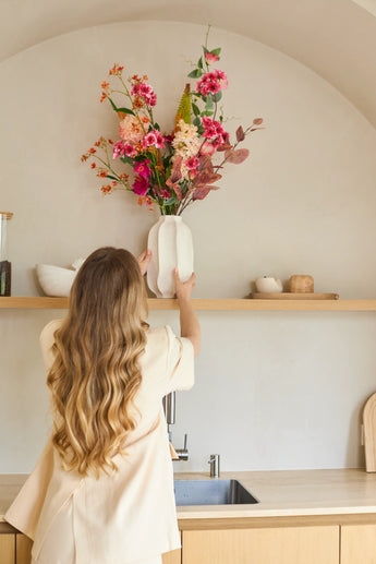 Une femme aux longs cheveux ondulés place un vase avec le bouquet de fleurs artificielles en soie Wild Peach sur une étagère de cuisine en bois au-dessus d'un évier. En arrière-plan, vous pouvez voir un design minimaliste dans des tons neutres.