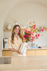 Une femme en tenue beige se tient dans une cuisine moderne. Elle sourit et tient le bouquet de fleurs artificielles en soie « Wild Peach », disposé dans un élégant vase blanc et rempli de fleurs colorées. Au fond se trouvent des étagères avec des ustensiles de cuisine et une cafetière.