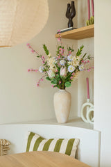Sur une étagère blanche se trouve un vase beige avec le bouquet coloré de fleurs artificielles en soie, « Lots of Love XL », à côté de bougies roses et d'objets décoratifs. Il y a un coussin rayé vert et blanc sur une chaise sous l'étagère.
