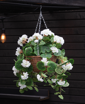 Le bégonia artificiel blanc dans un panier UV 70 cm, avec des feuilles vertes luxuriantes et des fleurs blanches, est suspendu à une chaîne en métal devant un mur en bois sombre avec une lampe lumineuse en arrière-plan.