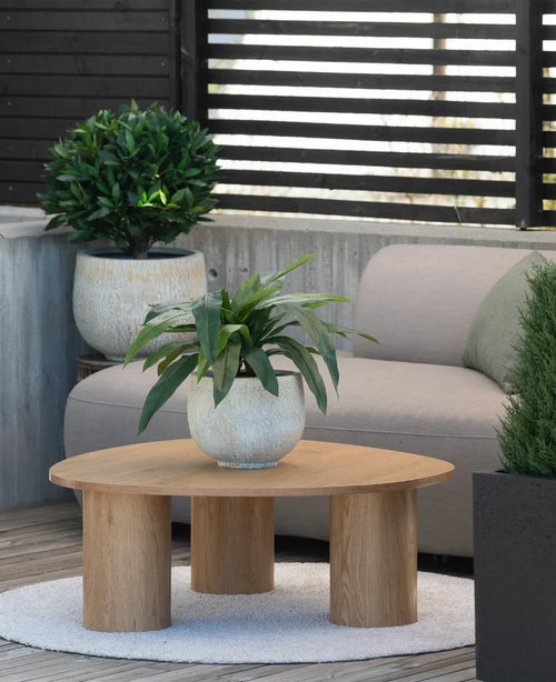 Sur un petit tapis à côté d'un canapé gris sur une terrasse se trouve une table basse en bois surmontée d'une plante artificielle Bird's Deluxe de 60 cm, avec quelques plantes en pot en arrière-plan.