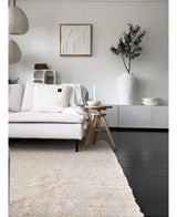 Un salon minimaliste avec un canapé de couleur claire, un tapis neutre, un buffet blanc, un olivier artificiel de 120 cm comme élément décoratif, une décoration murale abstraite et des tabourets en bois - le tout dans une douce ambiance méditerranéenne aux tons neutres.