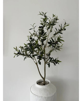 L'Olivier Artificiel 120 cm présente des feuilles vertes et des olives noires dans un pot blanc avec structure et galets, ce qui en fait un bel élément de décoration sur un mur uni gris clair.