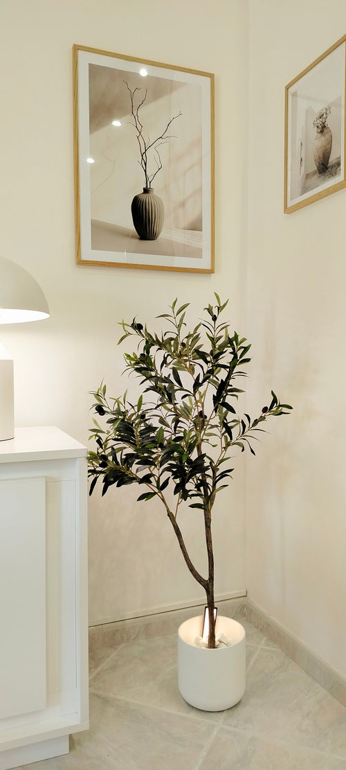 Un olivier artificiel de 120 cm aux feuilles vertes se dresse dans le coin d'une pièce lumineuse et ajoute une touche méditerranéenne. Au-dessus sont suspendues deux impressions d'art encadrées de vases avec des branches, tandis qu'un meuble blanc et une lampe sont partiellement visibles sur la gauche.