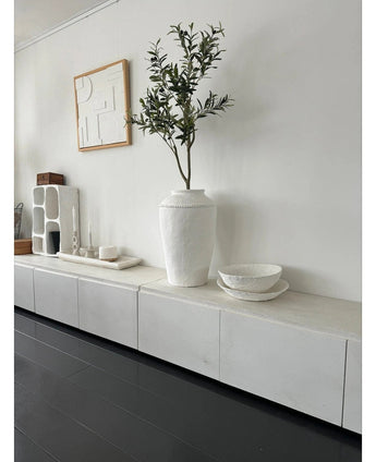 Une pièce minimaliste avec un buffet blanc, l'Olivier Artificiel 120 cm comme élément décoratif, des bols décoratifs blancs, une bougie et une décoration murale abstraite. Les parquets en bois sombre et les accents modernes créent une atmosphère méditerranéenne sereine.