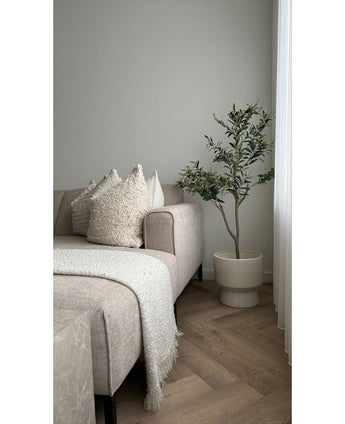 Un salon moderne avec un canapé beige, des coussins texturés, une couverture blanche à franges et un olivier artificiel de 120 cm comme élément décoratif à côté d'une fenêtre avec des rideaux blancs sur un sol clair à motif à chevrons.