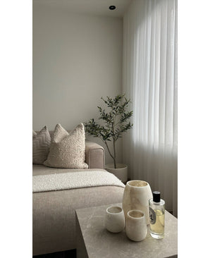 Un salon minimaliste dispose d'un canapé beige, de coussins texturés, d'un olivier artificiel de 120 cm comme décor, de rideaux blancs transparents et d'une table en marbre avec trois vases de couleur crème et du parfum. La lumière naturelle illumine l’espace tranquille.