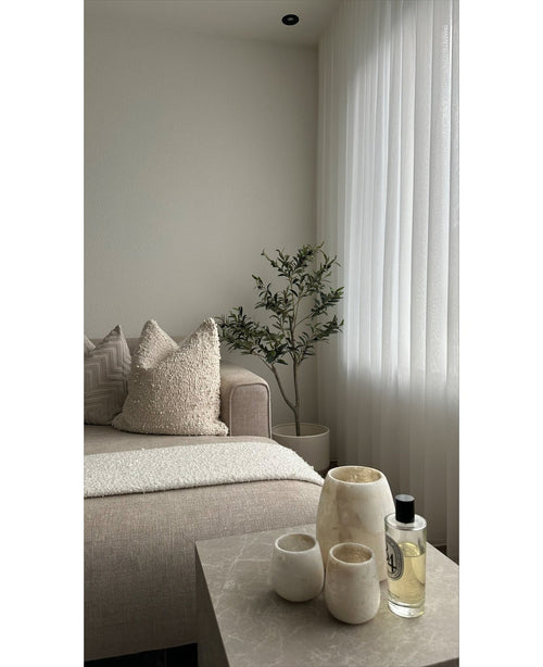 Un salon minimaliste dispose d'un canapé beige, de coussins texturés, d'un olivier artificiel de 120 cm comme décor, de rideaux blancs transparents et d'une table en marbre avec trois vases de couleur crème et du parfum. La lumière naturelle illumine l’espace tranquille.