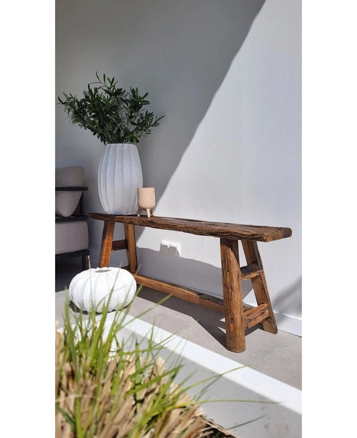 Un banc rustique en bois se dresse contre un mur blanc, à côté d'un grand vase blanc avec feuille d'olivier artificielle 90 cm UV pour une ambiance méditerranéenne. A proximité se trouvent un pouf rond blanc et un petit chandelier, avec la lumière du soleil projetant des ombres douces.