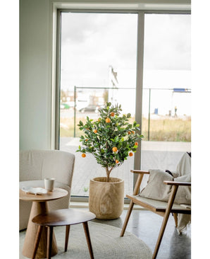 Un oranger artificiel de 120 cm se trouve près d'une grande fenêtre dans un salon moderne et lumineux avec une chaise beige, une table d'appoint en bois et une tasse à café sur une soucoupe.