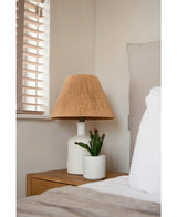 Une table de chevet avec une lampe blanche avec un abat-jour beige tressé et la plante succulente artificielle de 20 cm se trouve à côté d'un lit avec une literie blanche et beige, près d'une fenêtre avec des stores en bois.