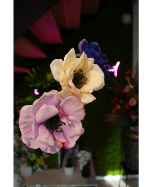 Gros plan de la fleur artificielle Anémone artificielle Real Touch violette 52 cm violet pastel et crème, sur fond d'éclairage vert et rose fluo, avec d'autres compositions florales visibles en arrière-plan.