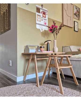Un coin salon moderne avec une table basse en bois clair avec la fleur artificielle Branches d'Anémone 56 cm rose dans un vase rose, accompagnée d'une lampe et de livres. À côté se trouve un canapé beige, rehaussé d’œuvres d’art murales et d’un tableau photo en forme de galerie sur le mur jaune.
