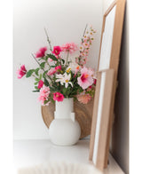 Une marguerite artificielle de 60 cm blanc/rose se dresse dans un vase blanc sur une surface blanche à côté d'une œuvre d'art dans un cadre en bois, avec un plateau rond en bois visible en arrière-plan.
