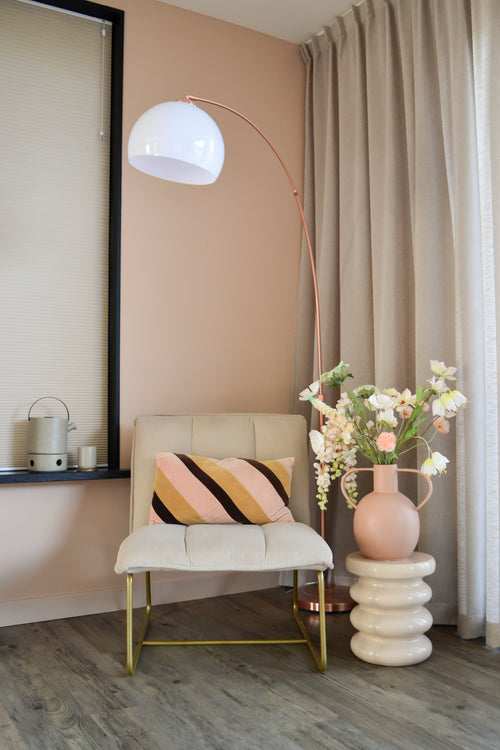 Un coin moderne et cosy avec une chaise couleur crème avec un coussin rayé, un lampadaire haut en forme d'arc, des rideaux beiges, un parquet et un vase rond avec des fleurs artificielles Fritillary 94 cm blanches sur une table d'appoint sculptée.