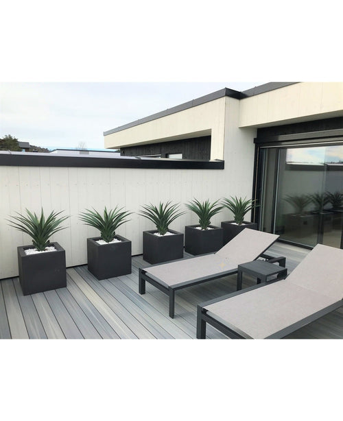 Un patio moderne comprend deux chaises longues grises et cinq jardinières carrées, chacune avec une plante artificielle Agava Pandora UV 85 cm, contre un mur lambrissé blanc et noir pour un look élégant et minimaliste.