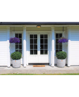 Des doubles portes blanches avec des panneaux de verre ornent une maison blanche à quatre colonnes. Deux topiaires de Buis Artificiel sur tige UV d60 cm et deux paniers suspendus à fleurs violettes décorent l'entrée, avec un paillasson devant les portes.