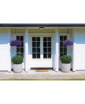 Des doubles portes blanches avec des panneaux de verre ornent une maison blanche à quatre colonnes. Deux topiaires de Buis Artificiel sur tige UV d60 cm et deux paniers suspendus à fleurs violettes décorent l'entrée, avec un paillasson devant les portes.