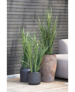 Trois plantes artificielles de 150 cm de gazon artificiel vert UV avec des feuilles longues et étroites se dressent sur une terrasse en bois à côté d'un canapé d'extérieur gris clair, contre un mur à lattes de bois sombre.
