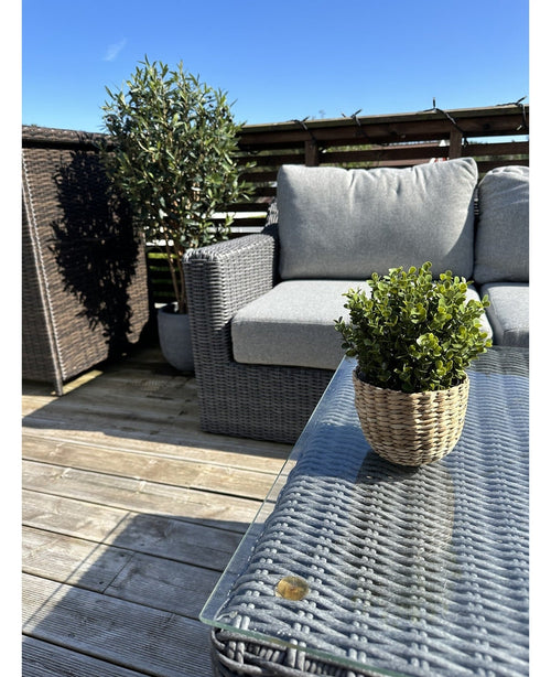 Une terrasse avec un banc et une table en osier gris, avec une plante verte de Buis Artificiel en pot UV 19 cm sur la table et une plante en pot plus grande à côté du banc, le tout sous un ciel bleu clair.