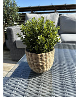 La Plante Artificielle Buxus vert en pot UV 19 cm, avec des feuilles vertes dans un pot en forme de panier en osier, se dresse sur une table d'extérieur en verre avec des meubles de patio et une balustrade en bois visible sous un ciel bleu clair.