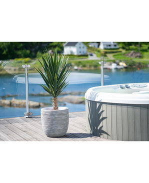 Une plante artificielle de yucca de 100 cm UV se dresse sur une terrasse en bois à côté d'un bain à remous avec balustrade en verre, surplombant un lac tranquille et des collines verdoyantes parsemées de maisons.