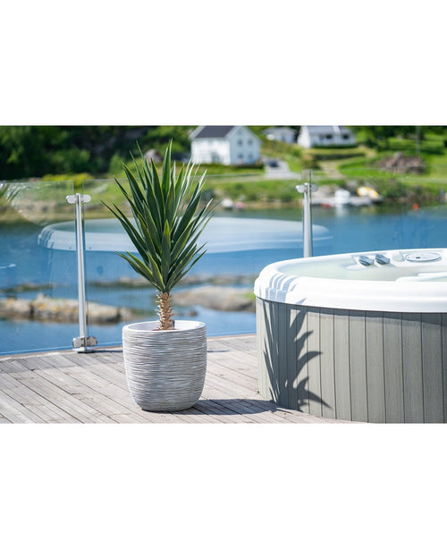 Une plante artificielle de yucca de 100 cm UV se dresse sur une terrasse en bois à côté d'un bain à remous avec balustrade en verre, surplombant un lac tranquille et des collines verdoyantes parsemées de maisons.